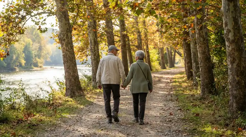 An older couple walking side by side on a tree-lined river trail in golden morning light, seen from behind